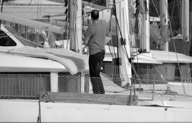 İtalya, Sicilya, Akdeniz, Marina di Ragusa (Ragusa Eyaleti), limandaki katamaran yelkenli teknesinde bir adam. 