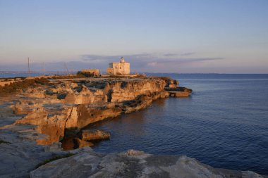 İtalya, Sicilya, Akdeniz, Egadi takımadaları, Favignana Adası (Trapani Eyaleti); gün batımında bir deniz feneri ve adanın kayalık sahili manzarası