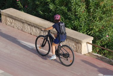 İtalya, Sicilya, Akdeniz, Marina di Ragusa (Ragusa Eyaleti), 19 Eylül 2024, deniz yolundaki bisiklet yolunda bisiklet süren kadın motosikletçi