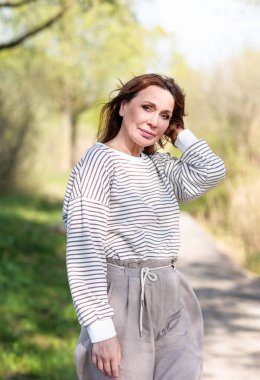 Güneşli bir bahar öğleden sonrasında orta yaşlı güzel bir kadın doğada poz veriyor. Güneş ışığı ve ağaçların gölgelerinin etkileşimi görsel bir etki yaratır..