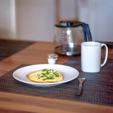 Bir fincan kahveyle servis edilen taze yeşil soğanlı yumuşak bir omletin yer aldığı basit ve lezzetli bir kahvaltı. Arka planda kahve demliği olan ahşap bir masanın üzerine kurulmuş rahat bir masa,