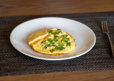 Taze soğanla kaplı mükemmel pişirilmiş bir omlet, temiz beyaz bir tabakta, tadını çıkarmaya hazır. Kabarık dokusu ve parlak garnitürüyle omlet basit ama tatmin edici bir şey sunuyor.