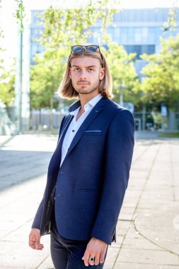 Uzun sarı saçlı, şık bir mavi takım elbiseli ve beyaz gömlekli genç bir adam modern mimari bir arka plana karşı güvenli bir şekilde açık havada poz veriyor. Aydınlık ve kentsel bir ortam..