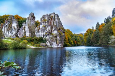 The Externsteine rock columns, standing 40 meters tall, form a striking natural monument in a peaceful nature reserve in Germany. Surrounded by autumnal colors, the reflection of the rocks and foliage create a picturesque scene on the lake. clipart