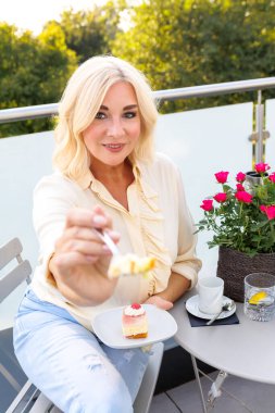 Sarışın kadın kahve, kek ve köpüklü suyla balkonda dinleniyor. Günlük kıyafet, güneşli gün, dostane atmosfer, çiçek süsleme. Yazın açık hava yaşam tarzı.