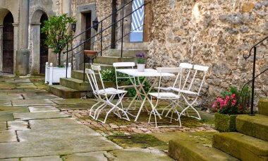 Büyüleyici taş bir avluda sandalyeli beyaz metal bahçe masası. Eski duvarlar, saksılar ve merdivenler kırsal cazibe katıyor. Rahat ve zamansız atmosferi olan huzurlu bir açık hava ortamı..