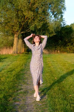 Uzun çiçekli elbiseli neşeli bir kadın doğanın tadını çıkarıyor, parkta yeşil bir yolda yürüyor ve dans ediyor. Ağaçlar ve yumuşak bahar güneşiyle çevrili özgür ve mutlu görünüyor..