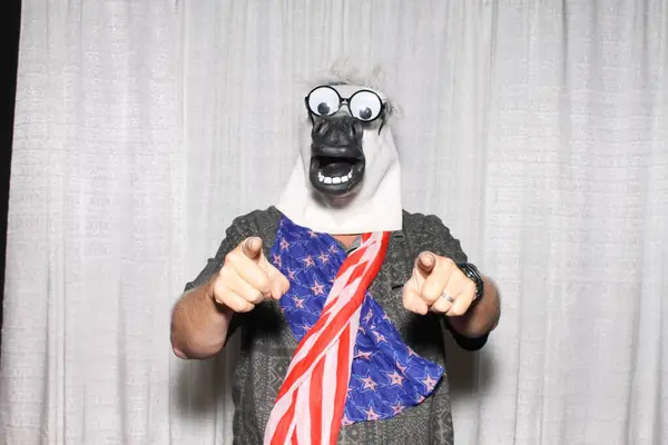 Unidentifiable person wears Horse Head Mask and scarf with american flag, poses while his pictures are taken in a Photo Booth.