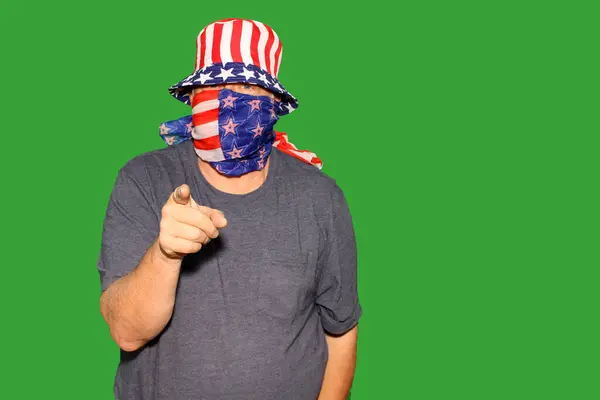 Green Screen. Chromakey. A man wears an American Flag Hat and Face Mask points at the camera while in front of a Chromakey aka Green Screen Wall. Green Screens are used for Special Effects and Editing