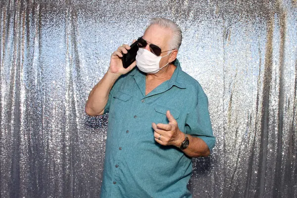 Photo Booth. A Caucasian Man wears a Anti-Covid 19 Face Mask as he talks on his phone and poses for photos in a Photo Booth with a silver sequin background.