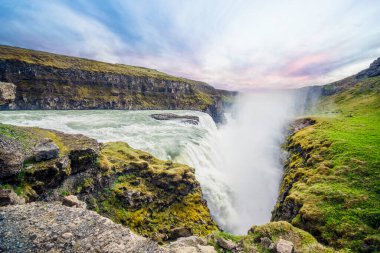 İzlanda şelalesi Gulfoss, gün batımında kayalıklarda yeşil yosunlarla çevrili.