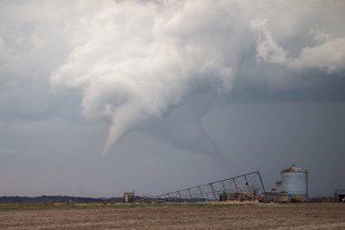 A white cone tornado hangs beneath a storm cloud over rural farmland with farm buildings and equipment in the foreground. clipart