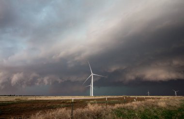 Wind turbines stand tall over a flat landscape with dark storm clouds looming on the horizon. clipart