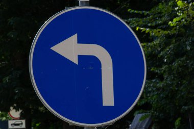 Trafik işaretleri, ağaçların bulanık arkaplanı ve parlak güneş ışığıyla sola dön.