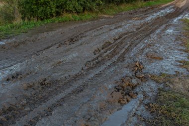 Vanishing dirt road with deep rut and puddles in village at sunset.