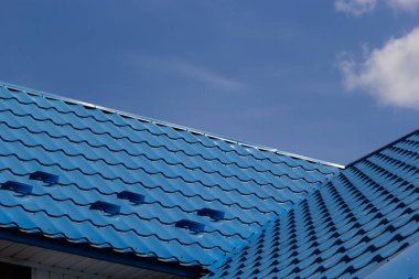 The roof of a house covered with sheets of blue metal tiles against the background of the sky on a summer day. Business selling building materials or repairing house roofs. clipart