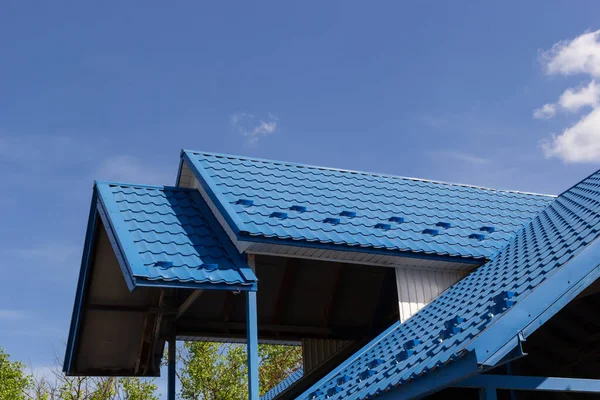 The roof of a house covered with sheets of blue metal tiles against the background of the sky on a summer day. Business selling building materials or repairing house roofs.