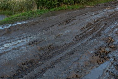 Vanishing dirt road with deep rut and puddles in village at sunset.