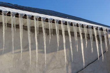 Icicles parlak mavi gökyüzüne karşı bir evin çatısında asılı. Evin çatısından sarkan buz saçakları olan bahar manzarası. Buz saçakları, kar şapkaları..