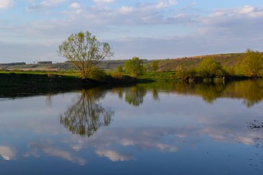 Dalgalı gökyüzü, beyaz bulutlar ve ağaçların simetrik olarak temiz mavi sulara yansıdığı sakin bir göl manzarası..