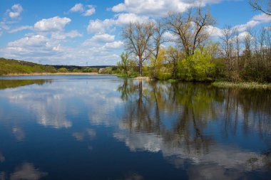 Dalgalı gökyüzü, beyaz bulutlar ve ağaçların simetrik olarak temiz mavi sulara yansıdığı sakin bir göl manzarası..