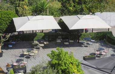 Gölgeli masalar ve sandalyeler açık mavi gökyüzünün altında yemyeşil ağaçlarla çevrili huzurlu bir açık hava yemek alanında düzenlenir..