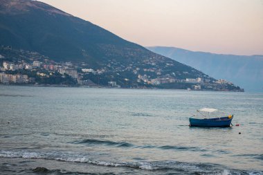 Akşamları, kıyı kasabası ve güneş batarken uzak dağlarla çevrili, sakin bir şekilde suda yüzen mavi bir tekne, sakin bir atmosfer yaratıyor..