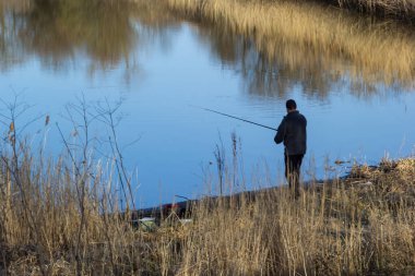 Huzurlu bir göletin kenarında elinde oltayla duran bir insan. Rüzgarda sallanan uzun otlar suyun yüzeyindeki yansımalar gibi barışçıl bir atmosfer yaratıyor..