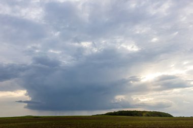 Karanlık bulutlar, arka planda küçük bir ormanlık alan olan yeşil bir alanın üzerinde oluşur ve akşam erken saatlerde karamsar bir atmosfer yaratır..