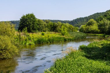 Sakin bir nehir, güneşli bir günde canlı yeşil çalıların ve ağaçların arasından süzülür. Çevreleyen tepeler ve berrak mavi gökyüzü manzaranın doğal güzelliğini arttırır..