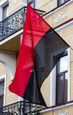 Canlı kırmızı ve siyah bir bayrak, sarı boyanmış binalar ve süslü demir parmaklıklarla dekoratif bir balkondan dalgalanıyor..