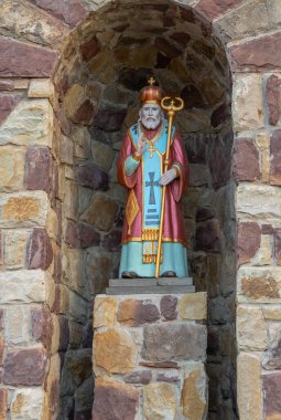 Dini bir figürün renkli heykeli taş bir oyukta duruyor, karmaşık detaylar ve canlı renkler sergiliyor, sıcak taş duvarların arka planına kurulmuş..