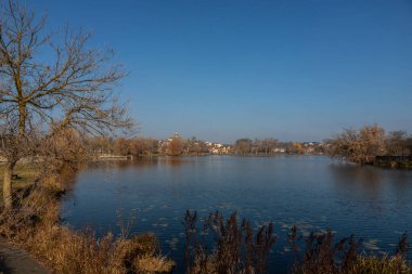 Sonbaharda ağaçlarla çevrili sakin bir nehir manzarası, açık mavi bir gökyüzü ve aşağıdaki huzurlu suya yansıyan uzak yapılar..