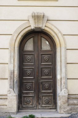 Ahşap çift kapı, geleneksel mimarinin zarafetini yansıtan, soluk taştan bir duvarın içine yerleştirilmiş süslü oymalar ve kavisli bir kemer içeriyor..