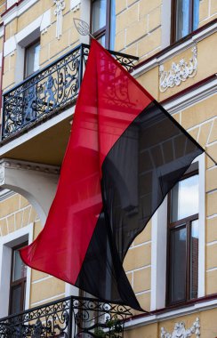 Büyük bir kırmızı ve siyah bayrak gururla eski bir binanın balkonundan dalgalanıyor canlı bir şehir ortamında açık mavi gökyüzüne karşı renkleri sergiliyor..