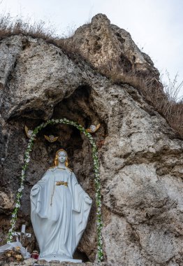 Beyaz giyinmiş bir kadın heykeli kayalık bir mağaranın girişinde duruyor, çiçeklerle ve kelebeklerle süslenmiş, doğada sükunet yayıyor..