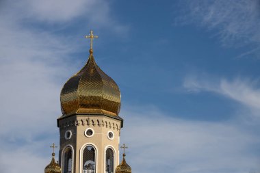 Güzelce işlenmiş bir kilise kulesinde altın bir kubbe ve detaylı pencereler bulunur. Görkemli bir şekilde açık mavi gökyüzüne yükselir. Arka planda yumuşak bulutlar vardır..