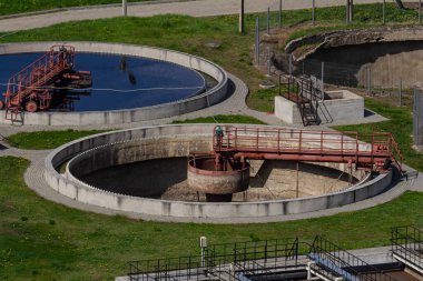 Dairesel atık su arıtma tankları aktif olarak bir tesiste su işliyor. Çevredeki yeşil alanlarda metal ekipmanlar ve bakım için kaldırımlar, verimli su yönetimi gösterileri.