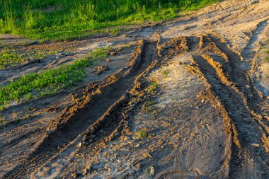 Lastik izleri toprak yüzeyde belirgin desenler oluşturur. Sıcak güneş ışığı manzarayı aydınlatırken yakındaki canlı yeşillikle çelişir..