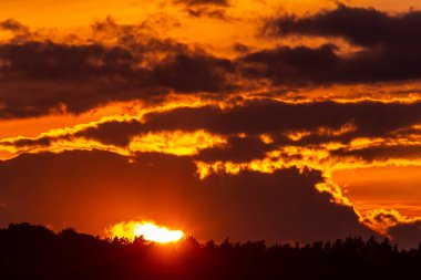 Güneş ufkun altına batarken, bulutları ve ağaç siluetlerini aydınlatırken, canlı günbatımı gökyüzünde sıcak renkler bırakır ve nefes kesici bir manzara yaratır..