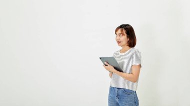 Kısa kahverengi saçlı, tişörtlü, kot pantolonlu, gülümseyen ve elinde dijital tabletle beyaz arka plan fotokopi alanının üzerinde duran çekici Asyalı genç bayan.