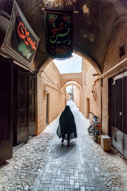 Yazd, İran - 26 Ekim 2018: Tarihsel Yazd şehrinin dar sokaklarında yürüyen siyah çarşaf giyen İranlı kadın. Antik şehrin eşsiz Pers mimarisi.