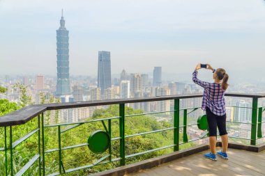 Genç, güzel bayan turist fotoğraf çekiyor ve dağın tepesinden Taipei manzarasının tadını çıkarıyor. Muhteşem Taipei silueti, Tayvan. Harika bir şehir manzarası. Gökdelenler ve şehir merkezindeki diğer modern binalar.
