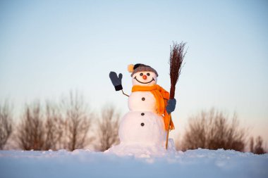 Funny snowman in knitted hat and yellow scalf with broom on snowy field. Blue sky on background
