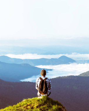 Bir turist, yazın pembe bir rhododendron çiçeği halısıyla kaplı bir uçurumun kenarında oturur. Arka planda sisli dağlar. Peyzaj fotoğrafçılığı
