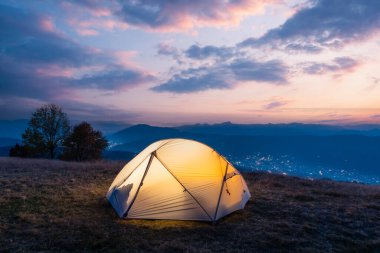 Gün batımında inanılmaz bir dağ manzarasının arka planında sıcak bir ışıkla aydınlanan turist çadırı.