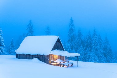 Karlı çam ağaçları ve kışın ormandaki bir dağ açıklığında yapayalnız bir ahşap ev. Noel kartpostalı. Karlı dağ ormanları