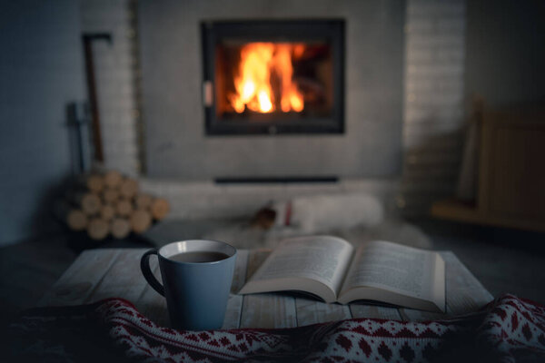 Cozy evening with reading near burning warm fireplace. Book and cup of tea on wooden table. Hygge and home coziness concept