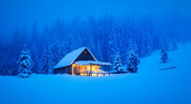 Ormanın göbeğinde, yapayalnız bir ahşap kulübe ve dağlık bir arazide karla kaplı çam ağaçları olan bir kış manzarası. Noel kartpostalı. Karlı dağ ormanları