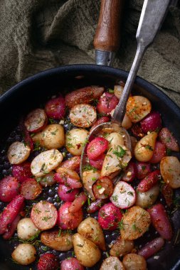 Fried radishes of different colors with herbs and spices on an iron frying pan with a spoon. Vegetarian food